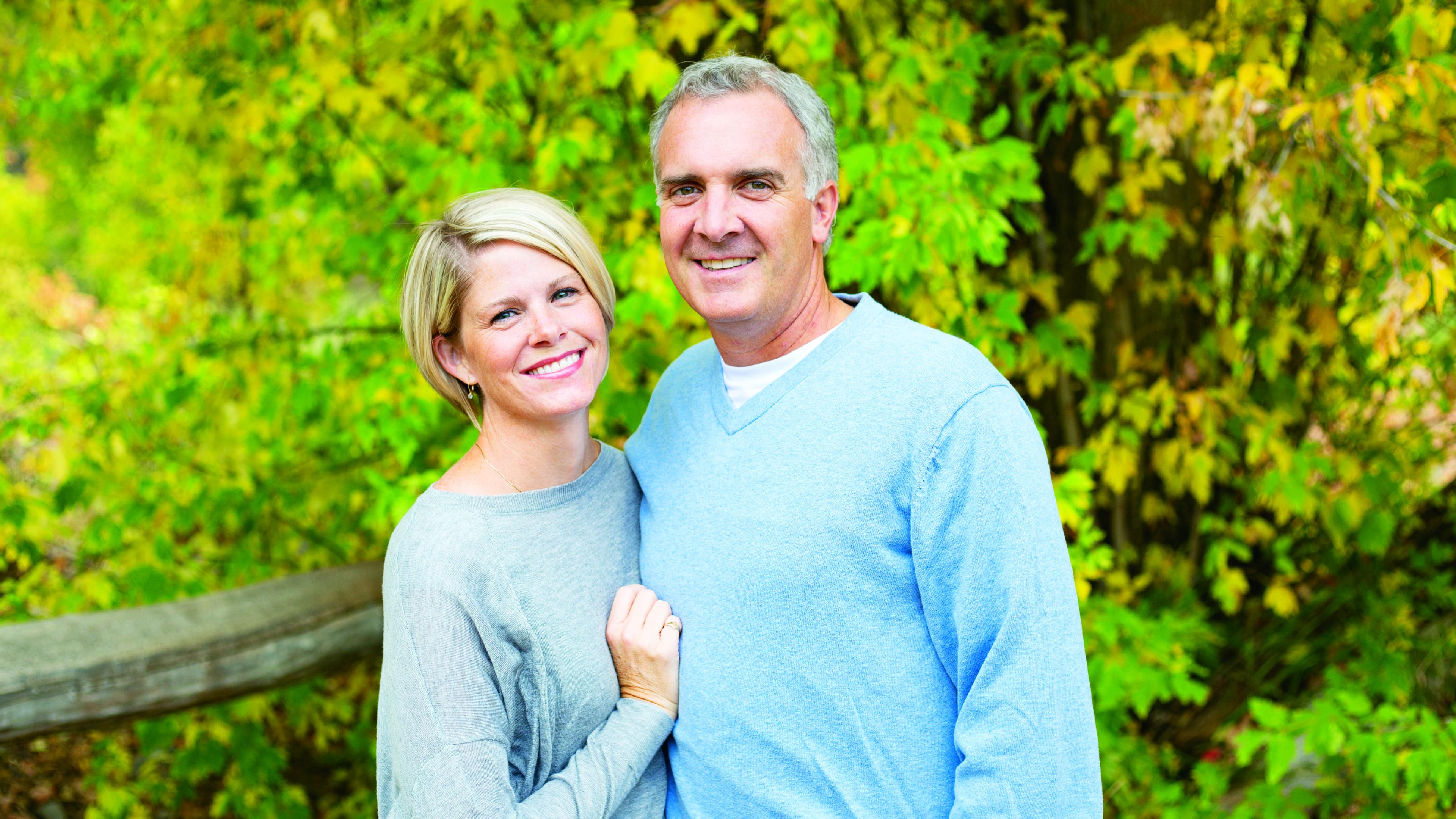 A smiling couple is standing in front of bright green bushes.